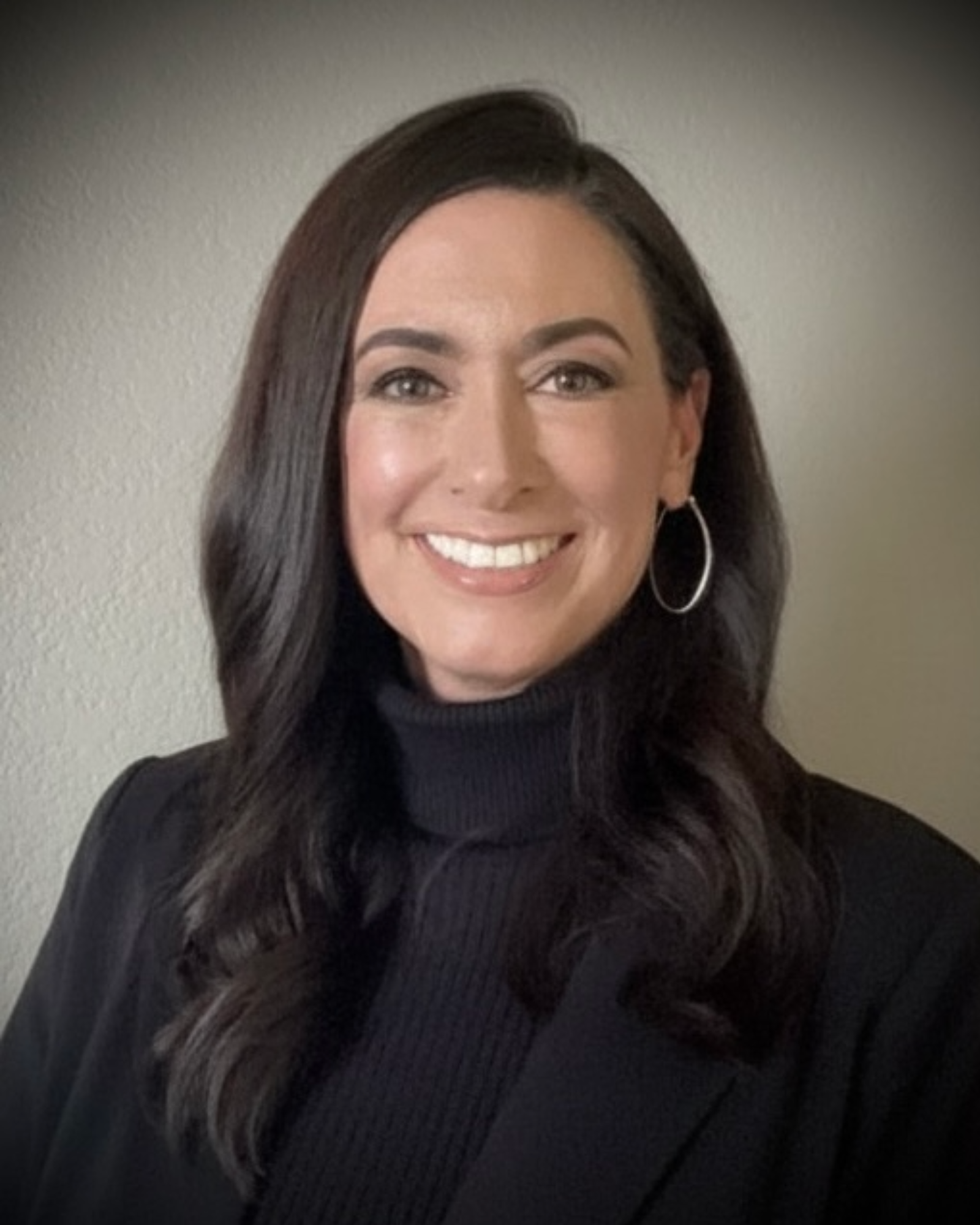 A professional headshot of a smiling woman with dark hair, wearing a black turtleneck and hoop earrings.